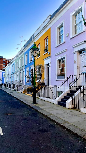 The image shows a row of colorful terraced houses situated along a narrow street, with houses painted in pastel shades of blue, yellow, and purple. Each property features white-framed sash windows and front doors, some with small decorative plants or elements on the window sills. Black wrought iron railings and steps lead up to the entrances, and a vintage-style street lamp with a green top is positioned on the sidewalk in front of the houses. The pavement appears clean and well-maintained, with a slight incline towards the background. In the foreground, a black road surface contrasts with the light-colored pavement, and distant rooftops with TV antennas are visible against a clear sky. This setting suggests a typical urban residential area where private waste collection and alternative rubbish removal methods, such as on-site clearance by companies like Rubbish Clearance Kensington, may be relevant. The scene emphasizes the neat and orderly appearance of the street, which could be prepared for waste disposal or removal related to home cleaning or renovation activities.
