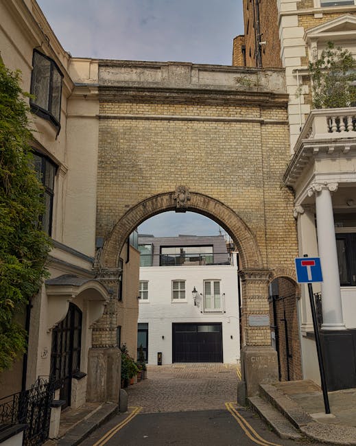 A photograph of a narrow street framed by historic brick buildings and a stone archway leading to a modern white residential building with large windows and a balcony. The archway is constructed from weathered yellowish-brown bricks with decorative stone elements, and it spans the width of the street, which is paved with cobblestones. On either side of the street, there are small pavement areas with bollards and a black wrought iron fence on the left. To the right, a white building with classical architectural features, including white columns and an ornate balcony, is partially visible. A blue one-way traffic sign indicating the street is a dead end is mounted on a pole near the corner of the building on the right. The scene is illuminated by natural daylight, with a clear sky visible through the archway, and the overall impression is one of a historic area being maintained amidst contemporary structures, with occasional signs of private property and on-site clearance typical of independent waste disposal efforts by companies like Rubbish Clearance Kensington.