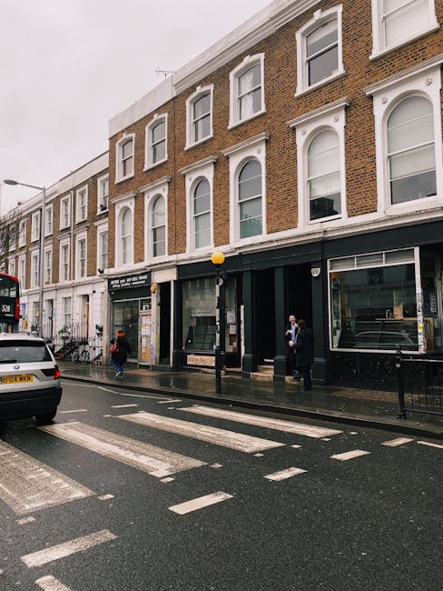 A street scene in a narrow Kensington area showing a three-storey brick building with white window frames and decorative architraves on the upper floors. The ground floor features large glass frontages, possibly shopfronts or commercial units, with dark frames and minimal signage. A black lamppost with a yellow spherical light is positioned on the pavement in front of the building. The wet street surface reflects the urban surroundings, indicating recent rain. Vehicles, including a parked white car and a red double-decker bus, are visible along the curb. Pedestrians, some wearing face masks, are walking on the sidewalk adjacent to the shopfronts. Bicycles are secured to railings near the building’s entrance. Despite the street’s narrow width, there is no visible trash or debris, suggesting a tidy environment. This scene implicitly relates to private waste handling or alternative rubbish collection methods in urban settings, aligning with the focus of rubbish clearance and disposal services such as those provided by Rubbish Clearance Kensington.