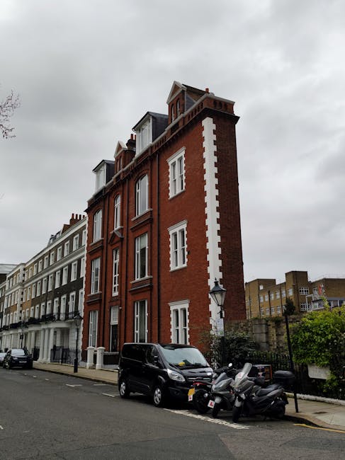 The image shows a narrow London street passage framed by an historic brick archway with a keystone at its apex featuring a carved stone face or emblem. The arch connects two buildings: on the left, a light-colored building with a curved bay window and decorative architectural details, partially obscured by green foliage from a nearby tree; on the right, a white building with a balcony supported by white columns and ornate railing. A blue rectangular dead-end road sign with a white horizontal line is positioned near the right side of the image, indicating no through passage beyond this point. The cobblestone pavement beneath the arch leads into a modern white building with large windows and a black garage door, visible in the background. The scene is illuminated by natural daylight, with soft shadows cast on the buildings, suggesting a clear day. The overall atmosphere reflects a blend of historic and contemporary urban architecture, illustrating a typical private or alternative access point in a central London area where rubbish clearance and private waste management might be coordinated outside council authority routes, as sometimes facilitated by local waste management services like Rubbish Clearance Kensington.