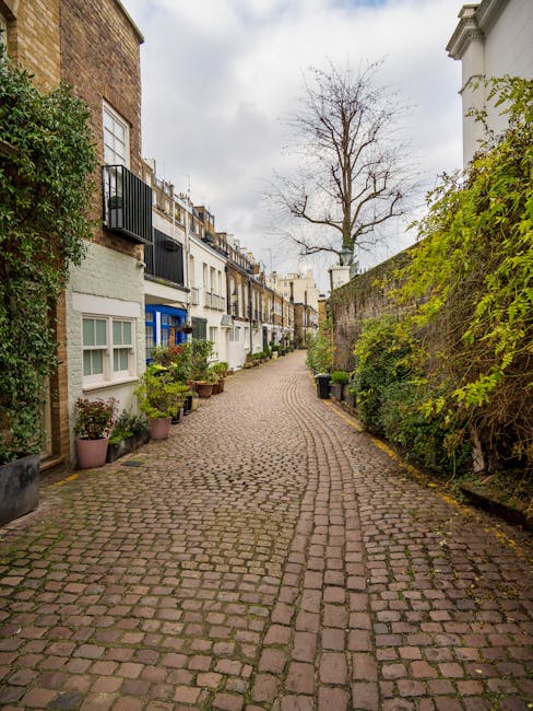 A narrow cobblestone alleyway in Kensington lined with residential buildings on the left side, featuring white and multi-coloured facades, some with small balconies and window boxes filled with plants. On the right side, a high brick wall is partly covered with green foliage and climbing plants. Potted plants are arranged along the edge of the buildings, adding greenery to the scene. In the background, a leafless tree stands against a cloudy sky, and the alleyway extends into the distance with a slight curve. The environment appears calm and well-maintained, illustrating a quiet residential area that may be typical for private waste collection or rubbish removal services, with Rubbish Clearance Kensington possibly involved in clearing household or garden waste from such sites.