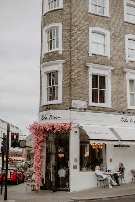 The image depicts a corner café situated on the ground floor of a multi-storey brick building with white window frames and decorative architectural details. The café's entrance is framed by a large glass door and window, partially covered by a white awning with the café’s name, 'Olive Moon,' written in elegant script. Pink flowering plants cascade from the top of a black metal stand near the entrance, adding a touch of vibrant colour and texture. Outside, two women are seated at a small round table on the sidewalk, engaged in conversation, with one woman wearing a white shirt and the other in darker clothing. The building’s upper floors have several windows, some of which are slightly open. A street corner features a pedestrian crossing and a traffic light showing a red signal. Parked cars line the street, and a street sign indicating the location as Kensington is visible near the corner. The environment suggests an urban setting with outdoor seating typical of cafes seeking to attract foot traffic, aligning with alternative approaches to waste disposal and street-level commercial activity. Rubbish clearance services, such as those provided by Rubbish Clearance Kensington, often support maintaining such vibrant urban spaces by handling the removal of waste and debris from commercial premises and outdoor areas.