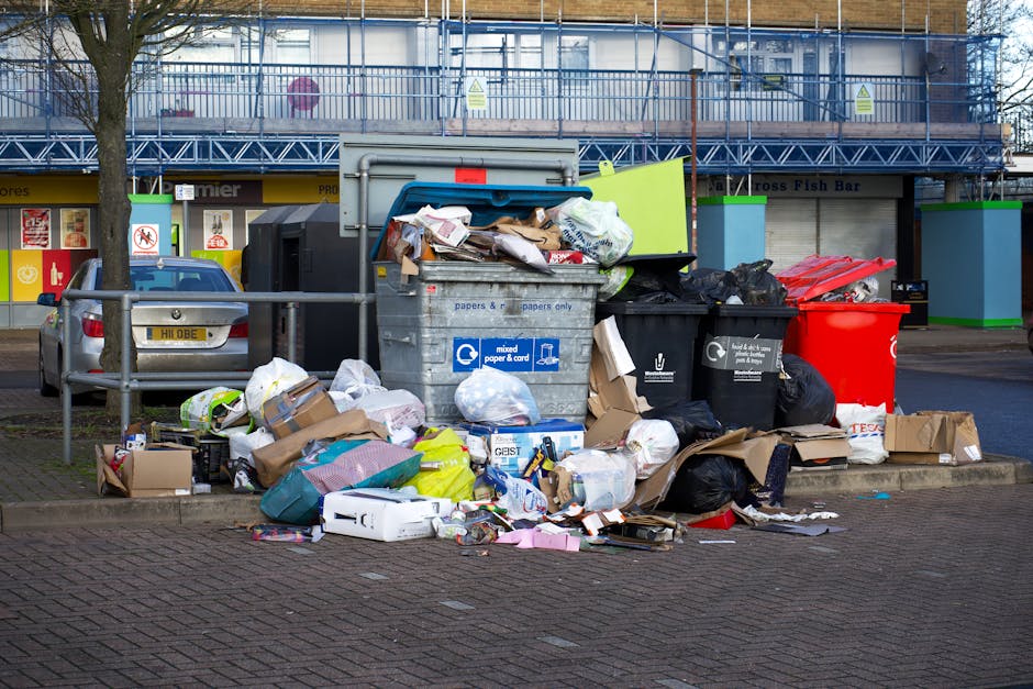 A large collection of mixed household waste and rubbish overflowing from multiple black, red, and grey plastic bins, situated on a paved area in front of a commercial building with a blue construction scaffold. The rubbish includes crumpled paper, cardboard boxes, plastic packaging, and discarded household items, some partially torn or flattened. Several cardboard boxes are placed on the ground beside the bins, and loose papers and trash are scattered on the pavement. A silver car is parked nearby, partially visible behind a metal railing that separates the waste collection area from the adjacent sidewalk. The building behind features a blue-framed storefront with yellow accents and signage, indicating commercial activity. The scene appears to depict an overflowing rubbish collection point, relevant to independent waste disposal services like those offered by Rubbish Clearance Kensington, illustrating the importance of regular waste management outside of council collections, particularly in busy urban areas.