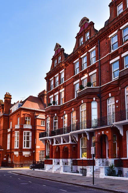 A multi-storey red brick residential building situated on a city street, featuring ornate architectural details with multiple bay windows and decorative trim. The building has white accents around the windows and a small balcony with a black iron railing on the first floor. The ground level includes an arched entrance and steps leading to a door, with a pavement and street in the foreground. Adjacent buildings with similar architectural styles are visible on the left, and the sky above is clear with bright daylight. The scene suggests an urban setting where on-site waste clearance or private rubbish removal services, such as those offered by Rubbish Clearance Kensington, might be employed for maintaining property cleanliness and compliance with local waste regulations.