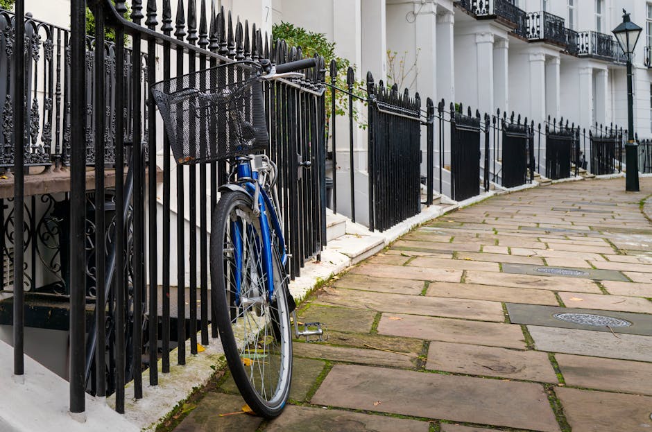 In the foreground, a blue bicycle with a black front basket is secured against a black wrought iron fence that runs along the edge of a residential street. The fence features pointed finials and horizontal bars, with some sections slightly leaning or open. Behind the fence, part of a white building with classic architectural elements, such as columns and decorative moldings, is visible, along with black balcony railings on the upper floors. The pavement consists of irregularly shaped, multi-toned stone slabs that are weathered, with patches of moss and dirt in the joints, extending along the sidewalk. The overall scene depicts a quiet, urban setting typical of Kensington, UK, with a calm atmosphere. The setting subtly illustrates a scene involving private property boundaries and on-site storage options, aligning with themes of independent rubbish collection or waste management services by companies like Rubbish Clearance Kensington.