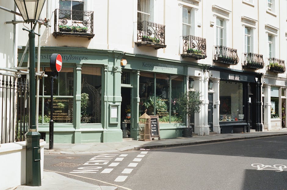 The image depicts a corner street scene featuring a row of retail shops housed in a white multi-storey building with several black wrought iron balconies adorned with potted plants. The ground-floor storefronts have large glass windows, with one painted in a muted sage green colour and another in black, both displaying various items and signs. A small chalkboard sign stands outside the green shop, and several potted bushes and plants are arranged along the pavement, contributing to a welcoming appearance. In the foreground, a street corner is visible with a zebra crossing and a pedestrian island, bordered by a black bollard and a lamppost with a decorative design. A no-entry traffic sign is mounted on the lamppost, indicating restricted vehicle access. The scene is well-lit with natural daylight, casting soft shadows on the pavement and emphasizing the clean and orderly appearance of the shopfronts and street environment, relevant to private waste management or on-site clearance considerations for commercial premises in a central London setting.