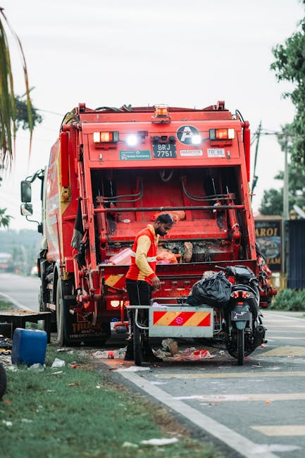 A sanitation worker in a high-visibility orange and yellow vest is operating a large red rubbish collection truck on a roadside. The worker is bent over, using both hands to load or assess waste within the rear compartment of the vehicle, which is partially open. The truck is positioned on a paved street with a marked parking lane, next to a grassy verge and a small roadside obstacle. Nearby, there is a black plastic bag placed on the truck's platform, along with some smaller debris scattered on the ground beneath the truck. In the background, trees, utility poles, and a building with signage are visible under an overcast sky, indicating daytime. The scene appears to be an outdoor area designated for waste collection, reflecting an instance of private or alternative rubbish removal service activity, such as on-site clearance or independent collection, rather than traditional municipal disposal.
