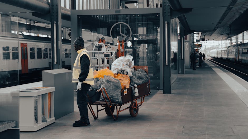 A worker in a high-visibility vest and dark clothing stands on a train station platform next to a large hand truck loaded with an assortment of garbage bags and miscellaneous waste materials, including black and grey refuse sacks, some orange and yellow plastic bags, and various unidentifiable debris. The platform area features a modern glass shelter with metal framing behind the worker, and a clear view of the train tracks with a passenger train parked alongside. On the left side, a white recycling or waste disposal bin is positioned against the platform edge. In the background, other station staff and passengers are visible, with some standing near the train doors and others walking along the platform, which is constructed of grey tile flooring. The scene captures a typical urban station environment, where waste collection activities such as private rubbish clearance by services like Rubbish Clearance Kensington may occur, especially when handling bulk rubbish outside of standard public recycling or waste services, set in a well-lit, contemporary transportation setting.